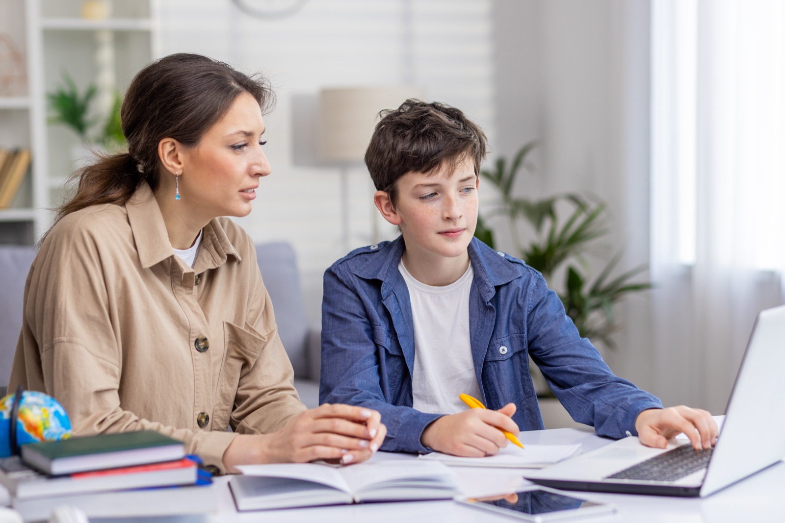 Mother and son studying together sitting at desk at home in living room, boy and woman tutor teaching at home, doing homework together.